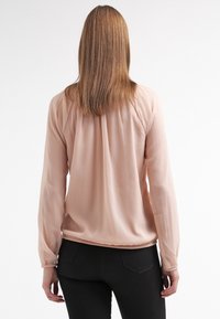 Světle růžová blůza s dlouhým rukávem, jemná textura, skladby na zádech a zaoblený lem. Nosit s černými přiléhavými kalhotami.