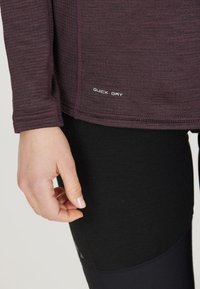 Mørkerød langærmet træningstop i struktureret stof med et "QUICK DRY" mærke ved kanten. Parret med sorte leggings.