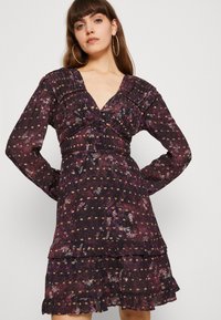 Vestido escuro com padrão floral, mangas compridas, decote em V, busto franzido e bainha com babados em camadas. Apresenta detalhes em roxo, rosa e dourado no padrão.