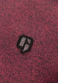 Close-up van rode gebreide stof met een klein geborduurd patch met een geometrisch ontwerp in zwart en grijs.
