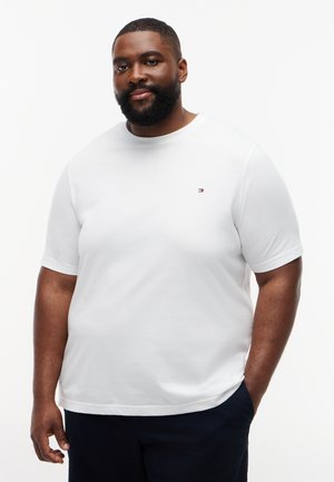 Bärtiger Mann, der ein weißes T-Shirt mit kurzen Ärmeln und kleinem Logo auf der Brust trägt, steht mit einer Hand in der Tasche vor einem schlichten Hintergrund.