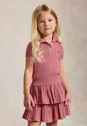 Mladé dievča s dlhými blond vlasmi má na sebe ružové polo šaty s krátkym rukávom a vrkočovým vzorom, s vrstvenou sukňou, stojí na neutrálnom pozadí.