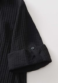Camicia nera, con texture e un design stropicciato. Presenta un risvolto con un accento di bottone scuro. Vista ravvicinata della manica.