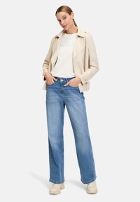 Trench beige, t-shirt bianco con scritte sottili, e jeans a gamba larga blu chiaro. Il modello indossa stivaletti beige. Design semplice e moderno.