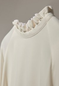 Blouse couleur crème avec un col texturé et froncé. Fabriquée en tissu lisse avec une coupe légèrement décontractée. Design minimaliste.