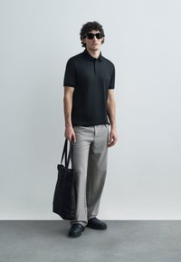 Black polo shirt, gray textured trousers, black tote bag, and black shoes. Model wears sunglasses and stands against a light background.