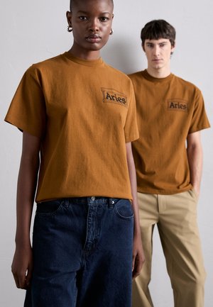 Braune Baumwoll-T-Shirts mit kurzen Ärmeln und einem Logo auf der Brust, kombiniert mit dunkelblauen Jeans und beigen Hosen. Glatte Textur.