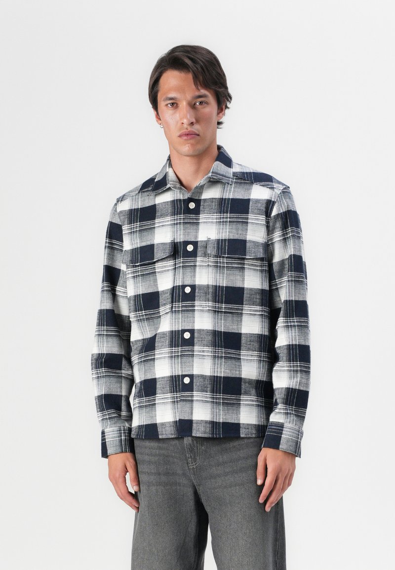 Marineblå og hvit rutete skjorte med lange ermer, knapper, brystlomme og løs passform. Stoffet virker mykt og middels tungt.