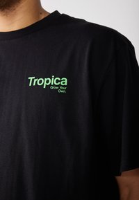 Svart bomull T-skjorte med grønn grafikk med teksten "Tropica" og "Grow Your Own" på venstre brystområde. Korte ermer.