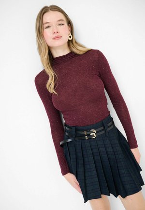 Top bordeaux a maniche lunghe con finitura testurizzata, abbinato a una gonna plissettata a quadri scuri con due cinture nere e dettaglio con fibbia dorata.