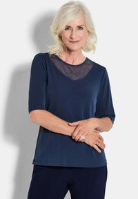 Top corto a maniche corte di colore blu navy con yoke in rete trasparente a pois. Tessuto morbido, vestibilità rilassata e scollatura arrotondata. Gli accenti includono una leggera lucentezza.