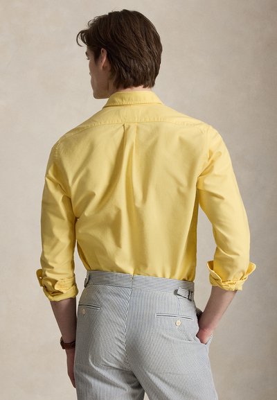 Homme vu de dos portant une chemise jaune à manches longues avec les poignets retroussés et un pantalon rayé gris clair avec des poches à l’arrière.
