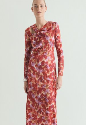 Femeie purtând o rochie lungă până la glezne, cu mâneci lungi, având un model abstract floral în culorile roșu, mov și maro și un design unic cu decupaj la umăr.