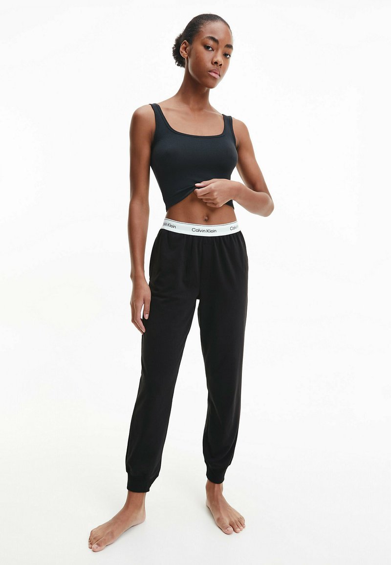 Schwarzes geripptes Tanktop und schwarze Jogginghose mit elastischen Bündchen und einem weißen Calvin Klein Logo-Bund. Keine Muster, minimalistisches Design.