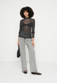 Top nero trasparente a maniche lunghe con texture a rete, abbinato a jeans grigi a gamba larga. La modella tiene in mano una piccola borsa nera e indossa ballerine con la punta appuntita.