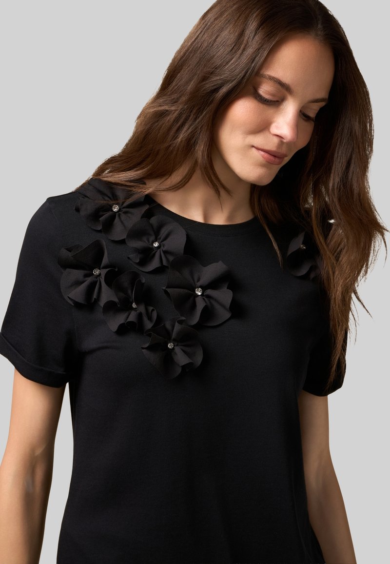 Camiseta de algodón negra con un racimo de flores de tela con acentos de cristal en el hombro izquierdo. Mangas cortas, cuello redondo.