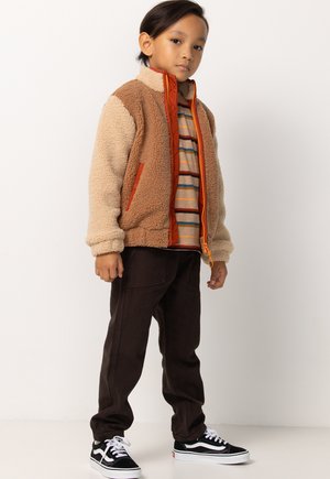 Fleece jack met beige en bruine secties, oranje rits, gestreepte shirt eronder, donkere broek en zwarte sneakers met witte accenten.