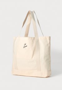 Lett stofftote i lys beige med doble håndtak. Har en logo i mørk trykk på den ene siden. Enkel, rektangulær design. Glatt tekstur.