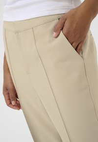 Pantalons beige ajustés avec une texture lisse, comportant des poches latérales et un design slim fit. Le tissu est léger et légèrement élastique.