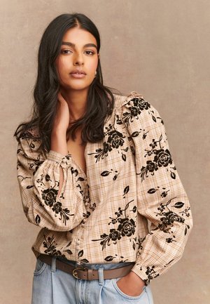 Vrouw met lang donker haar, gekleed in een beige check blouse met zwarte bloemenprint en lichtblauwe jeans, poserend met één hand op haar nek.