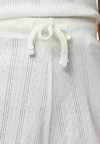 Tejido blanco con rayas verticales y pequeños patrones de diamante. Presenta una cintura con cordón y una textura suave.