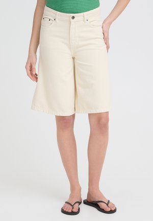 Vrouw draagt crèmekleurige shorts tot op de knieën, groene top en zwarte teenslippers, staand tegen een effen achtergrond.
