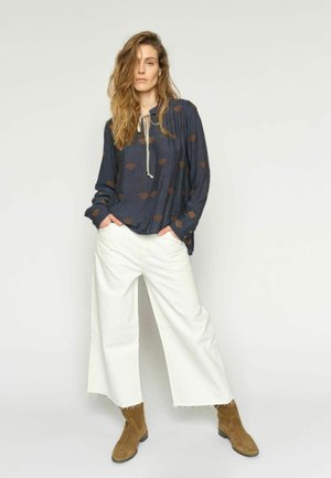 Naine pikkade laineliste juustega, kandes tumesinist mustrilist pluusi, laiu valgeid lühikesi pükse ja pruune seemisnahast pahkluu saapaid, seisab käed taskutes.