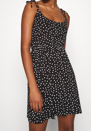 Femme portant une robe noire sans manches à pois blancs avec des bretelles nouées sur les épaules, se tenant devant un fond blanc uni.