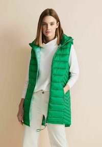 Zelená nadýchaná vesta s kapucí, s prošívaným designem, dvěma bočními kapsami a zapínáním na zip; nošena přes bílý svetr.