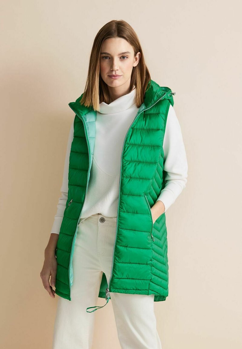 Zelená nadýchaná vesta s kapucí, s prošívaným designem, dvěma bočními kapsami a zapínáním na zip; nošena přes bílý svetr.