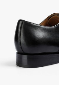 Zapato negro de cuero con acabado liso y pulido, tacón redondeado y detalle de costuras sutiles a lo largo del borde. Suela plana con ligera textura.