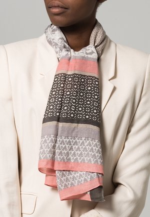 Gros plan d'une personne portant un blazer blanc et une écharpe à motifs dans des tons de rose, gris et noir, enroulée autour du cou.