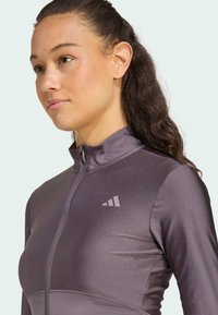 Dams zip-up jacka i en mörklila, glansig, atletisk tyg med hög krage och en subtil logotyp på vänster bröst.