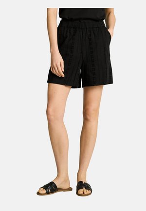 Frau trägt schwarze Shorts mit Ösenmuster, schwarzes Top und schwarze Slide-Sandalen, steht mit den Händen in den Taschen.