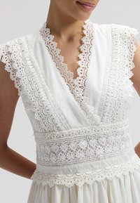 Γυναίκα που φοράει λευκό αμάνικο φόρεμα με διακοσμητικό δαντέλα με φλοράλ σχέδιο, V-λαιμόκοψη και διαφανές δαντελένιο ζωνάκι στη μέση.