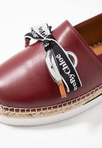 Chaussure en cuir bordeaux avec un lacet en ruban noir et blanc, œillet rond et plateforme en jute tissé. Elle est dotée d'une semelle en caoutchouc blanche.