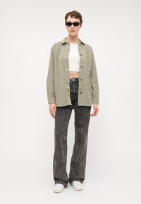 Chemise en velours côtelé vert clair, top blanc court, jeans foncés évasés à motif étoile, et baskets blanches ; accessoirisé avec des lunettes de soleil noires.