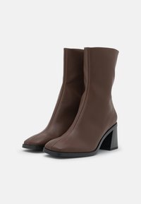 Pár hnědých kožených bot po lýtka s kulatými špičkami, černými blokovými podpatky a hladkým povrchem, vystavených na bílém pozadí.