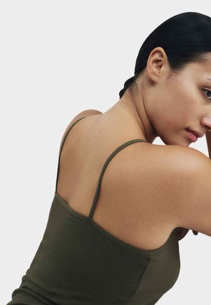 Kobieta o ciemnych włosach związanych z tyłu, ubrana w oliwkowozielony top na cienkich ramiączkach, widziana z tyłu i z boku na jednolitym jasnym tle.