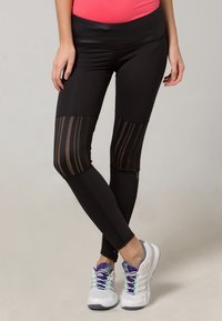 Czarne legginsy z przezroczystymi paskami na wysokości kolan, elastyczny materiał, wysoki stan, w połączeniu z białymi i fioletowymi butami sportowymi.