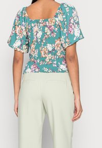 Blusa floreale in teal con maniche a sbuffo e scollatura quadrata. Presenta dettagli arricciati sul retro e un tessuto leggero e testurizzato.