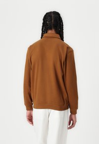 Brun sweatshirt med krage, långa ärmar och ribbade muddar. Slät textur, åtsittande fåll, bärs med vita byxor. Utsikt från baksidan.