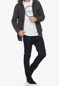 Cárdigan de punto gris con capucha sobre una camiseta blanca con un logo circular, combinado con jeans azul oscuro y zapatillas negras.