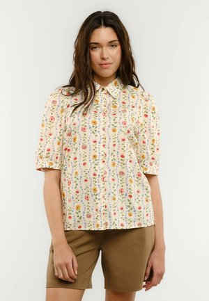 Kobieta z długimi ciemnymi włosami, ubrana w koszulę na guziki z krótkim rękawem z wielokolorowym pionowym wzorem kwiatowym oraz brązowe krótkie spodenki sięgające do kolan.