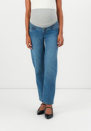 Personne enceinte portant un jean de maternité bleu avec une bande ventrale élastique grise et des ballerines noires Mary Jane, debout sur un fond blanc.