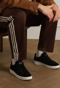 Schwarze Wildleder-Slipper mit einer weißen Gummisohle. Tragen Sie sie zu braunen Sporthosen mit weißen seitlichen Streifen und auf einem strukturierten Holzboden.