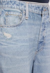 Jean bleu clair taille haute avec texture délavée, poche avant, passants pour ceinture, fermeture par boutons, et petite zone usée sur la cuisse.