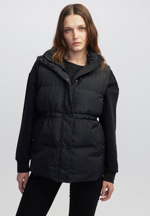 Vrouw met schouderlang bruin haar in een zwart gewatteerd mouwloos vest over een zwart longsleeve-top tegen een effen achtergrond.