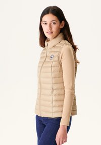 Beige isolierte Weste mit einem hohen Kragen, gesteppter Textur, vertikalen Nähten und einem runden Logopatch auf der Brust. Getragen über einem Langarmshirt.