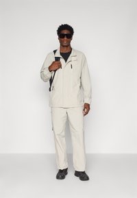 Light beige, waterproof jacket with a zip front, side pockets, and a collar, paired with matching trousers and black shoes.